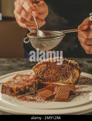 Weibliche Hände halten ein Sieb hoch, aus dem Kakaopulver auf Schokoladenfelder und ein Schokoladen- und Erdnussdessert fällt Stockfoto