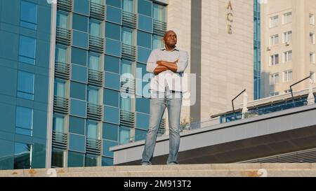 Ethnischer afroamerikanischer älterer männlicher Tourist-Geschäftsmann mittleren Alters mit gekreuzten Armen, der in der Stadt draußen auf einem blauen modernen Büro steht Stockfoto
