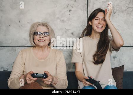 Seniorenmutter und Dudson spielen gemeinsam zu Hause Videospiele, sitzen und halten den Gamecontroller und haben Spaß. Aktivitätsbeziehung. Familienhaus Stockfoto