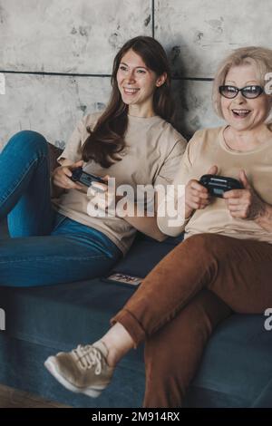 Erwachsene Tochter mit ihrer leitenden Mutter, die zu Hause Videospiele mit Konsolensteuerung spielt und auf dem Sofa sitzt. Aktivitätsbeziehung. Familienheim Freizeit Stockfoto