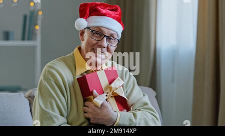 Ein alter weißer, älterer 60s-Mann mit Brille, männlich, der Weihnachten Weihnachtsmann-Red-hat trägt, Großvater, Großvater, Großvater, auf der Couch zu Hause, umarmt das Weihnachtsgeschenk-Paket Stockfoto