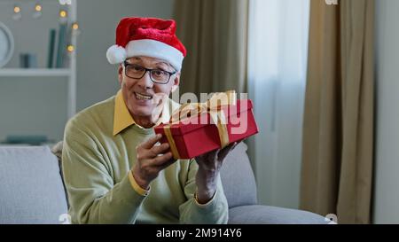 Alter weißer, älterer Großvater im Weihnachtsmann-Hut zu Hause, der ein Lied singt, das tanzt, sich bewegt, Spaß hat, Weihnachten Neujahr zu feiern Stockfoto