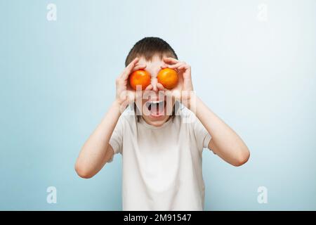 Glücklicher lächelnder Junge mit zwei Mandarinen, die seine Augen auf blauem Hintergrund verdecken Stockfoto