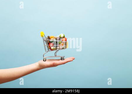 Handgehaltener Einkaufswagen mit Obst und Gemüse auf blauem Hintergrund. Konzept für den Einkauf von Lebensmitteln im Internet Stockfoto