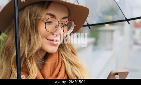Eine kaukasische lächelnde Frau aus nächster Nähe unter dem Schirm mit einem Telefon im Freien, eine Frau, die ihr Smartphone mit Hut und Brille hält und sich mit dem Handy unterhält Stockfoto
