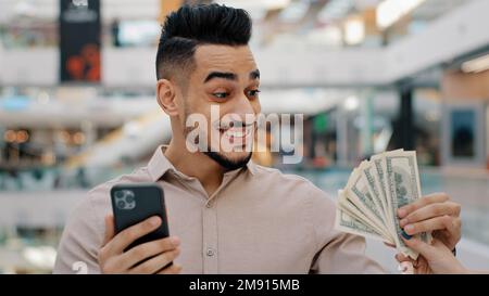 Schockiert überraschter, ethnisch indischer Sieger männlicher Kerl, der mit dem Handy surft, online wettet Casino spielen mit Smartphone, leicht Geld erhalten, verblüfft Stockfoto