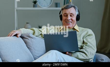 Aufgeregter, glücklicher alter Mann, der sich mit Laptop und Kopfhörern einen Film ansieht, der auf einer bequemen Couch liegt und den älteren Großvater lächelt, während er sich entspannt Comedy ansieht Stockfoto
