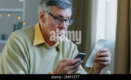 Kaukasischer alter 60s-jähriger Großvater, der ein Smartphone mit einer mobilen E-Bank-Anwendung in der Hand hält. Rechnungsbudget mit Papier berechnen Stockfoto