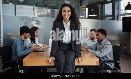 Zufriedene, lächelnde weibliche Führungskraft Chefin Geschäftsfrau, die auf einem Tisch sitzt, im Büro lächelt vor dem Hintergrund multirassistischer Kollegen Stockfoto