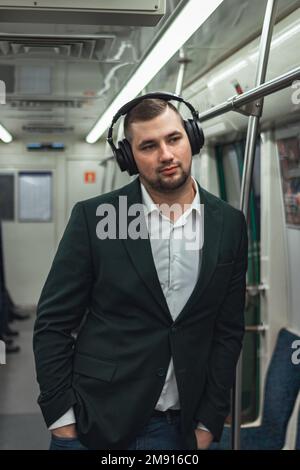 Mann junger weißer Passagier eine Person steht positiv fröhlich, hört Musik über Kopfhörer, lächelt und fährt morgens mit der U-Bahn. Konzeptabonnement für Musik. Stockfoto