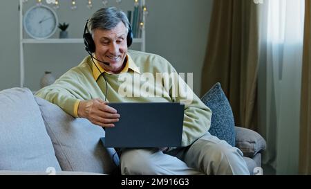 Ein erwachsener, sprechender 60s-jähriger Mann, der weit von zu Hause entfernt arbeitet, sitzt im Headset auf dem Sofa und spricht mit einem Webcam-Laptop. Älterer 60s-Online-Lehrer-Coach Stockfoto