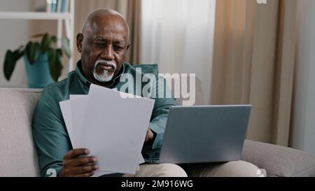Afroamerikanischer Geschäftsmann mit Papierkram. Ältere 60s-jährige, glatzköpfige Unternehmer, die auf einem Laptop tippen, verwalten die Finanzen des Haushalts zu Hause Stockfoto