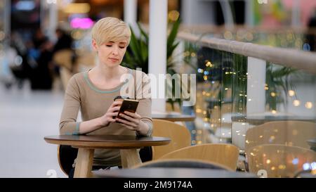 Erwachsene mittleren Alters 40s Weiße Geschäftsfrau, die an einem Café-Tisch sitzt, Kaffee trinkt, Tee trinkt und auf dem Handy telefoniert und sich mit dem Smartphone unterhält Stockfoto