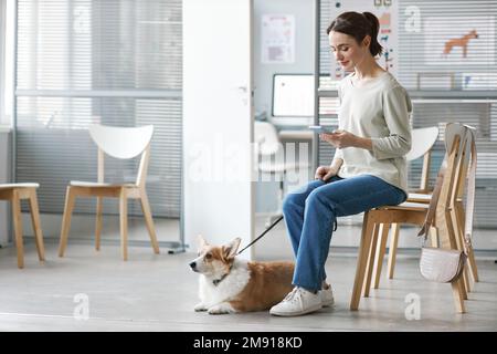 Junge brünette Frau in Freizeitkleidung, sitzend auf einem Stuhl, mit Handy und der Leine ihres Hundetieres, während sie auf einen Tierarzt in Kliniken wartet Stockfoto