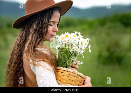 Eine Frau mittleren Alters in einem weißen Kleid und einem braunen Hut hält einen großen Strauß Gänseblümchen in den Händen. Wildflowers für Glückwünsche Stockfoto