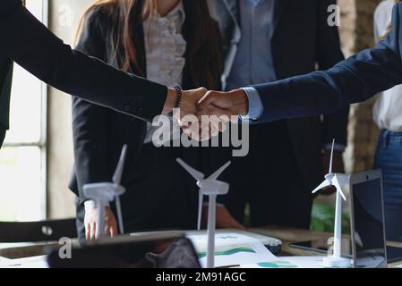 Handschlag zwischen zwei Geschäftspartnern, umgeben von Modellen und Diagrammen von Windkraftanlagen. Ideal zur Veranschaulichung von Themen wie Partnerschaft, erneuerbare Energien, Stockfoto