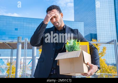 Ein afroamerikanischer Geschäftsmann wurde mit einem Karton gefeuert, in dem Büromaterial im Freien unglücklich war. Stockfoto
