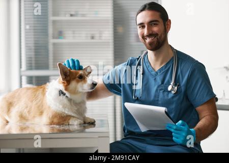Glücklicher junger Tierarzt mit Stethoskop am Hals und medizinischem Dokument in der Hand, der bei der Untersuchung des Corgi-Hundes in die Kamera schaut Stockfoto
