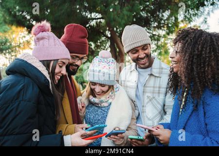 Eine Gruppe von multiethnischen jungen Freunden mit Mänteln und Hüten, die lächeln und die sozialen Medien mit einer Handy-App ansehen. Porträt von glücklichen Menschen, die Spaß beim Teilen von Medien haben und mit einem Smartphone lachen. Kommunikationskonzept. Hochwertiges Foto Stockfoto