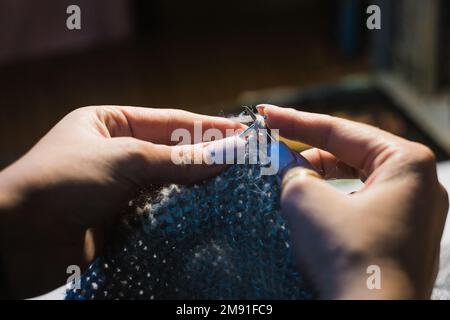 Nahaufnahmen von Händen, die das Kleid aus hellblauer Wolle auf verschwommenem Hintergrund stricken. Mit kreisförmigen Nadeln Stockfoto