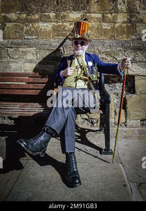 Porträt eines stilvollen Mannes, der auf einer Bank sitzt. Er trägt futuristische Steampunk-Kleidung im Retro-Stil und einen Hut aus Holz. Stockfoto