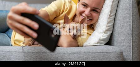 Junge brünette Frau, die Selfie mit ihrem Smartphone macht, während sie sich mit dem kleinen Hund auf dem Sofa hinlegt. Stockfoto
