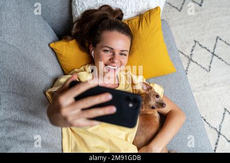 Junge brünette Frau, die Selfie mit ihrem Smartphone macht, während sie sich mit dem kleinen Hund auf dem Sofa hinlegt. Stockfoto