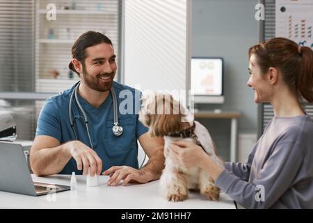 Glücklicher junger männlicher Tierarzt in Uniform, der eine Klientin mit ihrem Haustier ansieht, während er sie konsultiert und Medikamente für kranke Tiere verschreibt Stockfoto