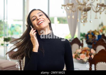 Goiania, Goiás, Brasilien – 10. Januar 2023: Eine glückliche junge Frau, die ein schwarzes langärmeliges Oberteil trägt, ihr Haar zurückwirft und mit ihrem Handy spricht. Stockfoto