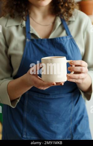 Eine Kunsthandwerkerin, die man nicht wiedererkennt, hält einen handgemachten Keramikbecher in der Hand. Kleine Unternehmen. Stockfoto