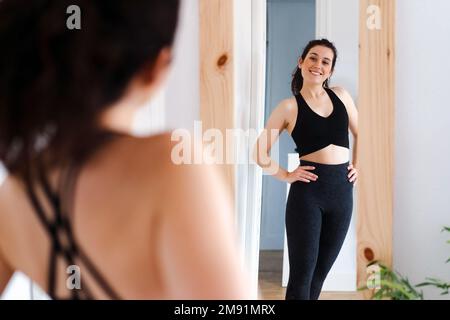 Junge, glückliche, schöne Frau, die in den Spiegel schaut, zufrieden mit den Ergebnissen ihres Trainings Stockfoto