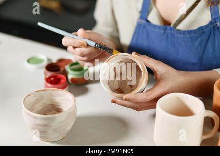 Frauenhände, Töpferstudio und Maler-Becher in der Werkstatt für Skulpturen, kreative Fertigung oder Design. Maler-, Keramikprodukte- und Bürstenverfahren, Stockfoto