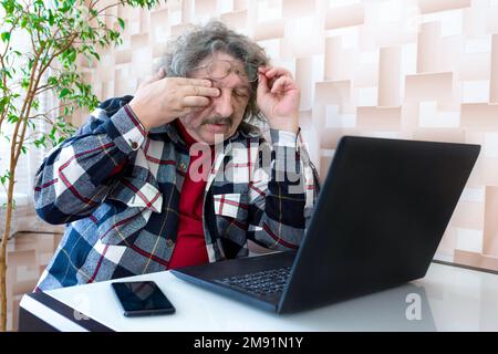 Ein älterer Mann, der seine Brille abnimmt und nach langer Arbeit am Computer unter Müdigkeit und Augenschmerzen leidet Stockfoto