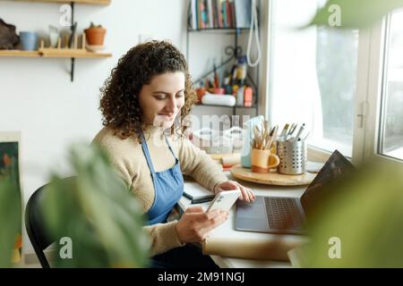 Eine Keramikfrau liest eine SMS auf ihrem Smartphone. Kreative Geschäftsfrau, die ein Geschäft mit einer Sammlung von Keramikprodukten leitet. Junger Entrepr Stockfoto