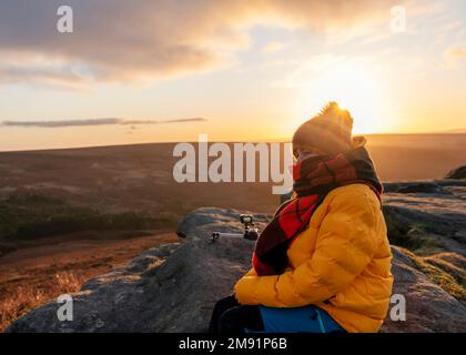 Frau in gelbem Mantel und orangefarbenem Schal erreicht das Ziel und sitzt auf dem Hügel oder Berg und nimmt bei Sonnenaufgang ein Video auf. Britische Kälte Stockfoto