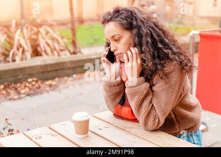 Verärgerte Frau mit langen Haaren in orangefarbenem Schal und Jacke, die im Café mit einer Tasse Kaffee sitzt. Stockfoto