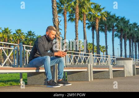 Ein Mann mittleren Alters mit hinterem Haar und Bart sitzt auf einer Bank in der Nähe von Palmengassen in Stadttypen und schickt Nachrichten an sein Handy Stockfoto