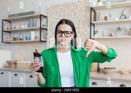 Unzufriedene junge Frau zu Hause in der Küche, die ein Stück Schokolade in der Hand hält. Mit der anderen Hand zeigt er auf einen Finger der Abneigung. Hasst Schokolade, Süßigkeiten, unterstützt eine Diät, zählt Kalorien. Stockfoto