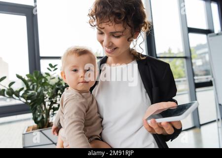 Kleines Baby sieht in die Kamera neben einer lächelnden Mutter mit Handy im Büro, Stockbild Stockfoto