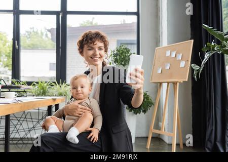 Freudige Geschäftsfrau im schwarzen Anzug, die mit dem Baby sitzt und Selfie auf dem Handy im Büro macht, Börsenbild Stockfoto