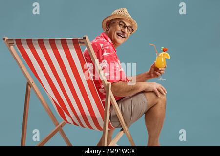 Reifer männlicher Tourist, der auf einem Stuhl sitzt und lächelt und einen Cocktail auf blauem Hintergrund hält Stockfoto