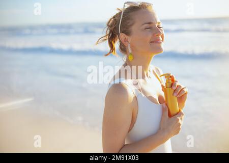Lächelnde stilvolle 40-jährige Frau in weißem Badeanzug mit Sonnencreme am Strand. Stockfoto