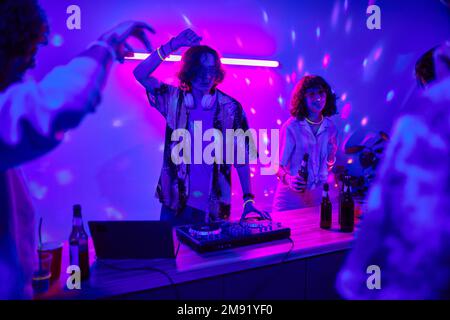 Junger DJ, der Musik auf dem Plattenspieler kreiert und tanzt, während er neben seiner Freundin mit einer Flasche Bier auf der Heimparty steht Stockfoto