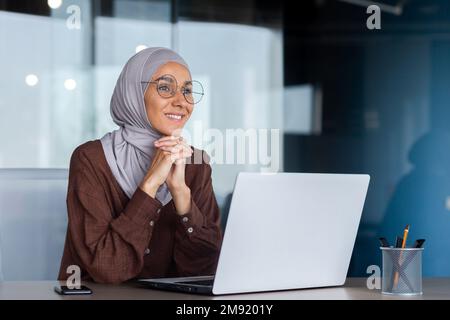 Lächelnde und träumige Geschäftsfrau, die im Büro mit einem Laptop arbeitet, Frau im Hijab und Brille, Büroangestellte, die mit der Arbeit am Schreibtisch zufrieden und zufrieden ist. Stockfoto