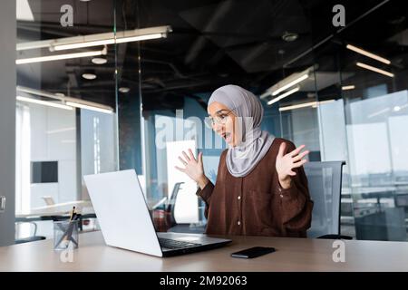 Erfolgreiche Geschäftsfrau in Hijab, die den Sieg und das Erreichen erfolgreicher Ergebnisse feiert, Frau, die auf den Laptop-Bildschirm schaut und die Hände in Win- und Triumph-Geste hochhält. Stockfoto