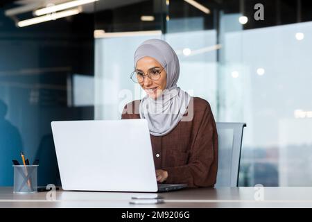 Lächelnde und träumige Geschäftsfrau, die im Büro mit einem Laptop arbeitet, Frau im Hijab und Brille, Büroangestellte, die mit der Arbeit am Schreibtisch zufrieden und zufrieden ist. Stockfoto