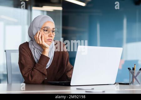 Ernste gelangweilte Geschäftsfrau im Büro, muslimische Frau im Hidschab, die denkt, während sie am Arbeitsplatz mit einem Laptop sitzt, Frau bei der Arbeit, die über Entscheidungen nachdenkt. Stockfoto