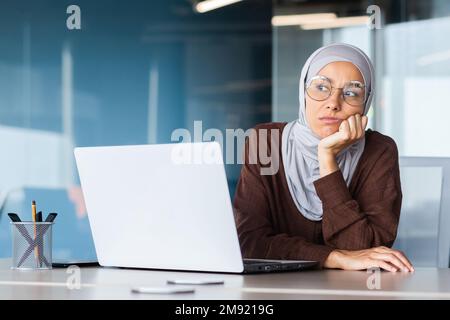 Ernste gelangweilte Geschäftsfrau im Büro, muslimische Frau im Hidschab, die denkt, während sie am Arbeitsplatz mit einem Laptop sitzt, Frau bei der Arbeit, die über Entscheidungen nachdenkt. Stockfoto