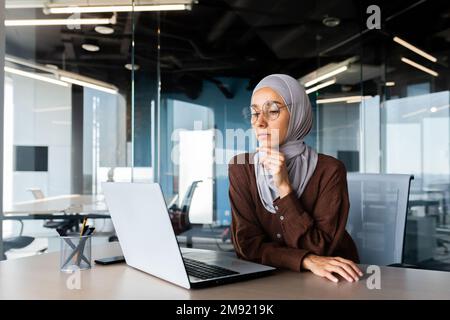 Ernste gelangweilte Geschäftsfrau im Büro, muslimische Frau im Hidschab, die denkt, während sie am Arbeitsplatz mit einem Laptop sitzt, Frau bei der Arbeit, die über Entscheidungen nachdenkt. Stockfoto