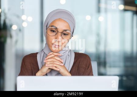 Ernste gelangweilte Geschäftsfrau im Büro, muslimische Frau im Hidschab, die denkt, während sie am Arbeitsplatz mit einem Laptop sitzt, Frau bei der Arbeit, die über Entscheidungen nachdenkt. Stockfoto